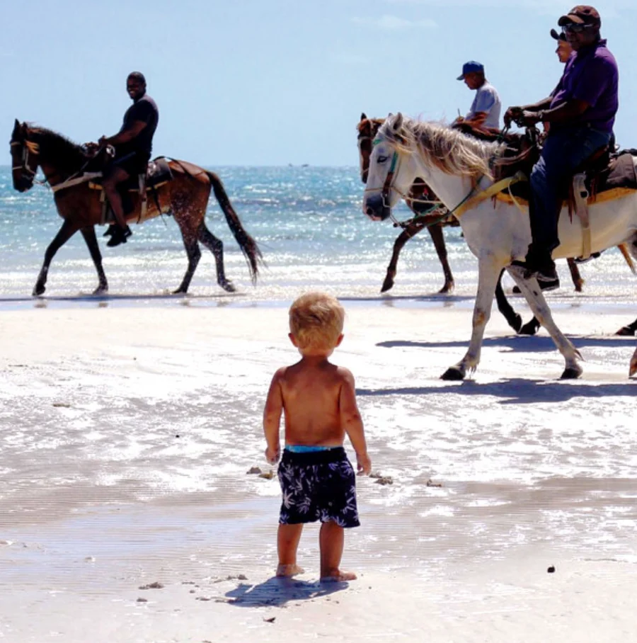 Child watching horseback riders on a sunny beach for family travel storytelling.